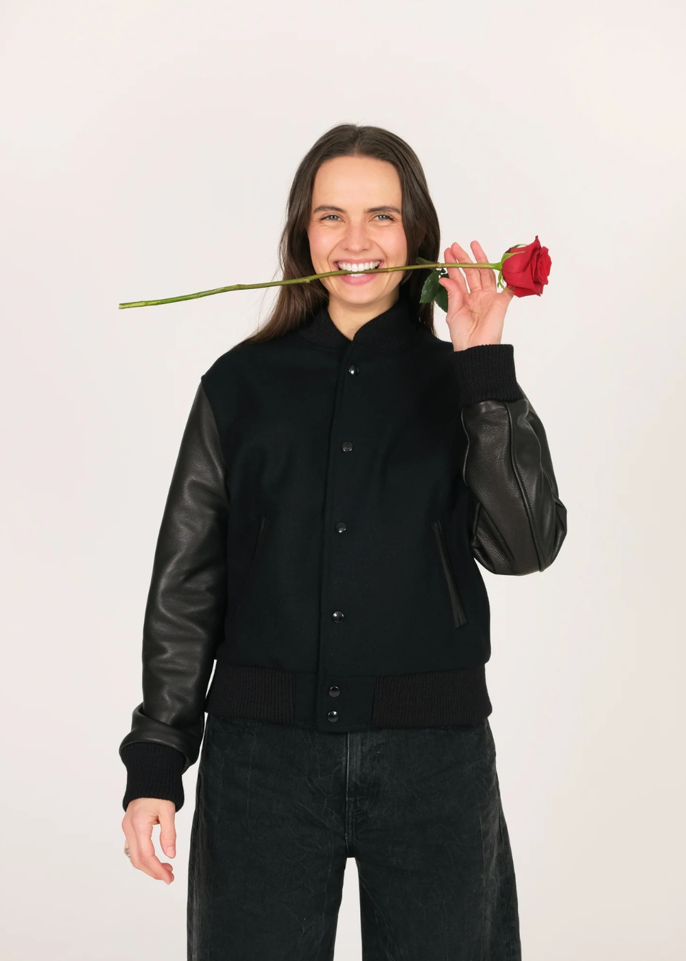 Woman wearing a stylish black varsity jacket with premium wool blend body and genuine leather sleeves, holding a red rose, perfect for a modern and classic look.